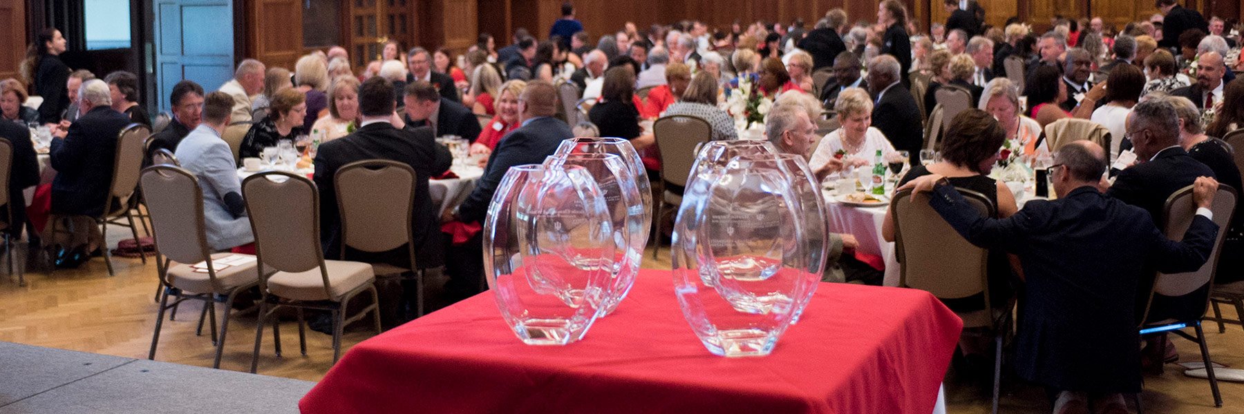 Four glass awards sit on a red table in front of a crowd of people sitting at round tables