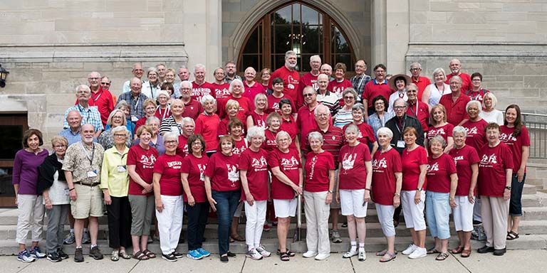 A large group of people wearing red and white stand on the steps of a limestone building with their arms in the air