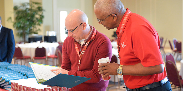 Two men in red look at a page in a blue binder