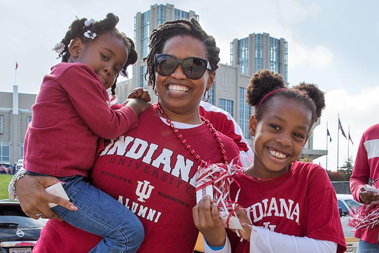 An African American woman wearing an IU shirt smiles while holding a small child and standing next to another.