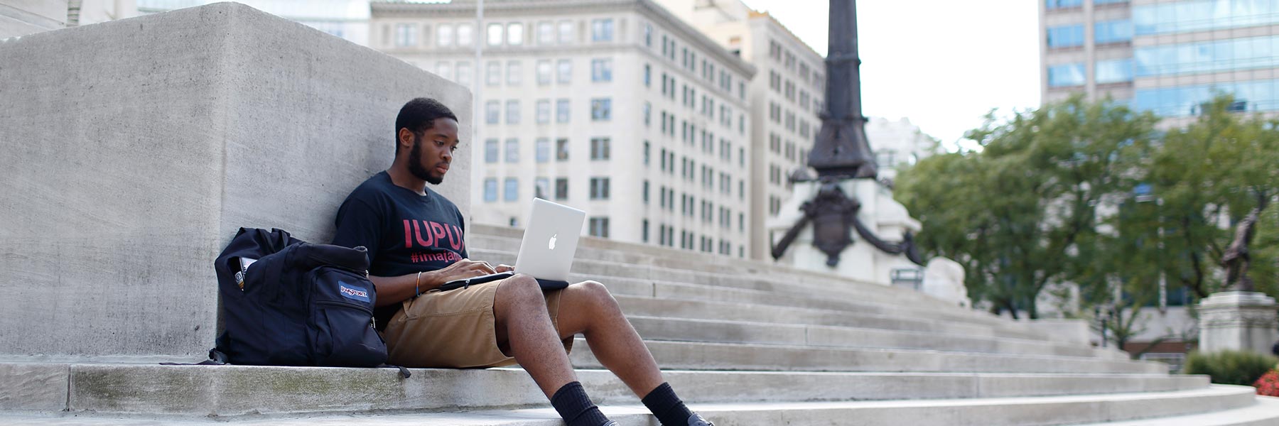 An African American man in a black IUPUI T-shirt sits on cement steps with a computer