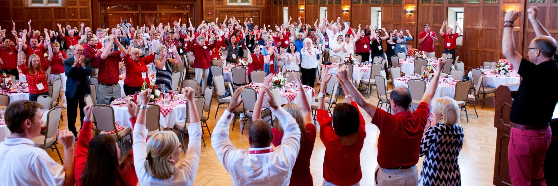 A large group of alumni stand in a circle in a wood paneled room doing the fist and blades.
